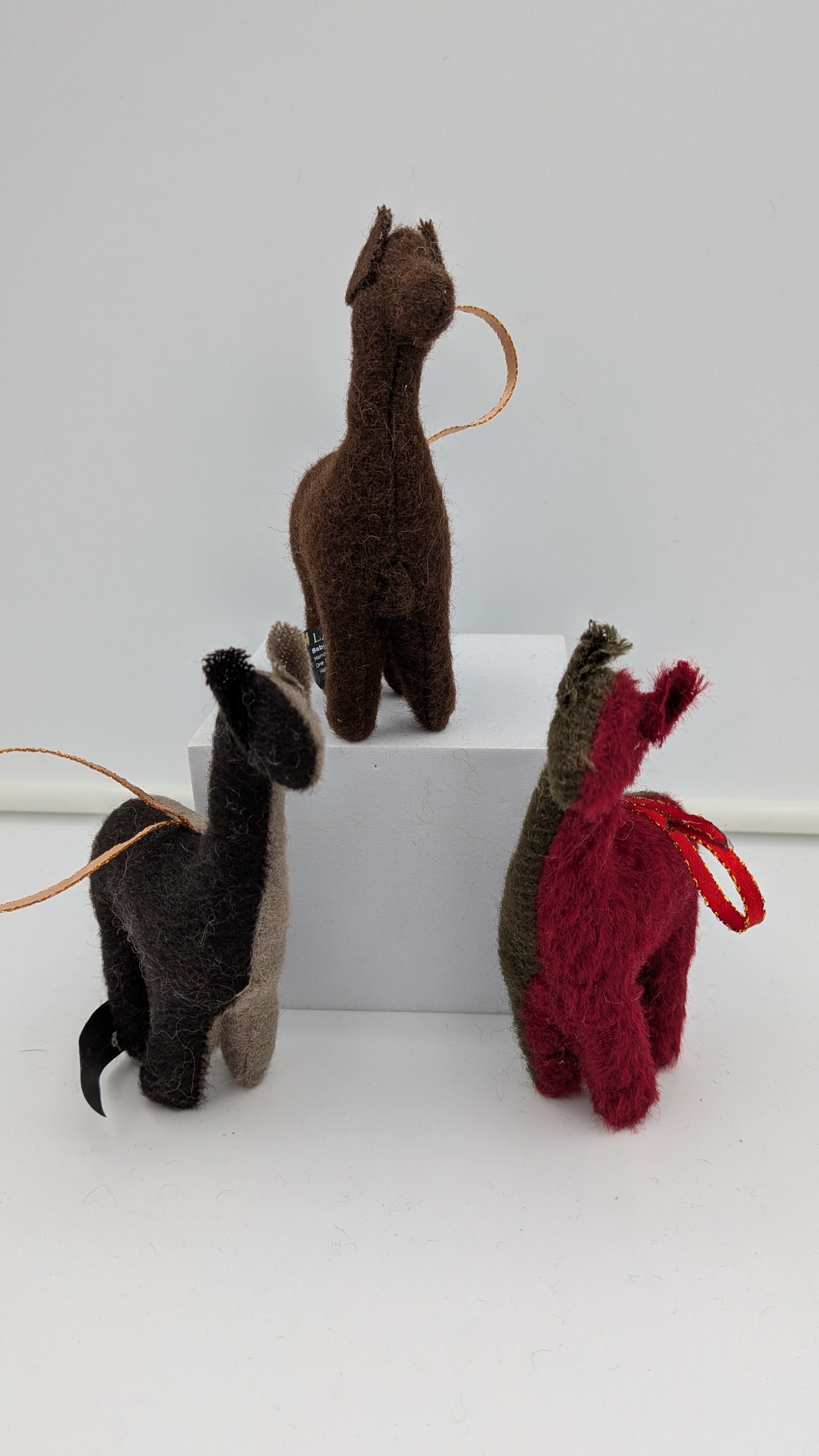 Three small felt llama or alpaca-shaped ornaments in brown, black, and red, each with a looped string and hanging loop, displayed on a white block against a light gray background.