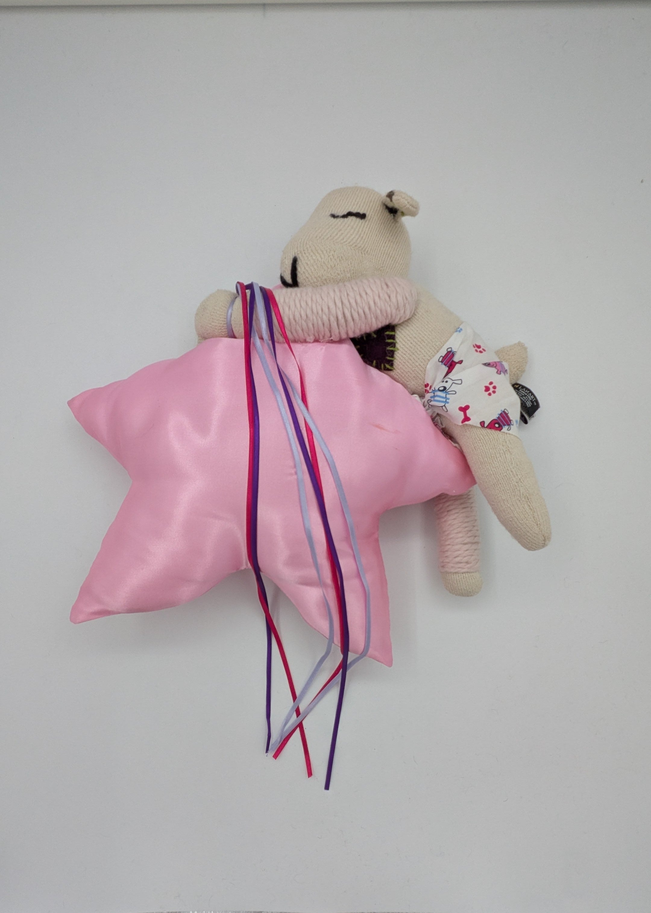 A small beige plush toy or stuffed animal lying on a white surface, wrapped in a shiny pink satin cloth with long multicolored ribbons tied around it. The toy has embroidered facial features and a tiny floral-patterned accessory.