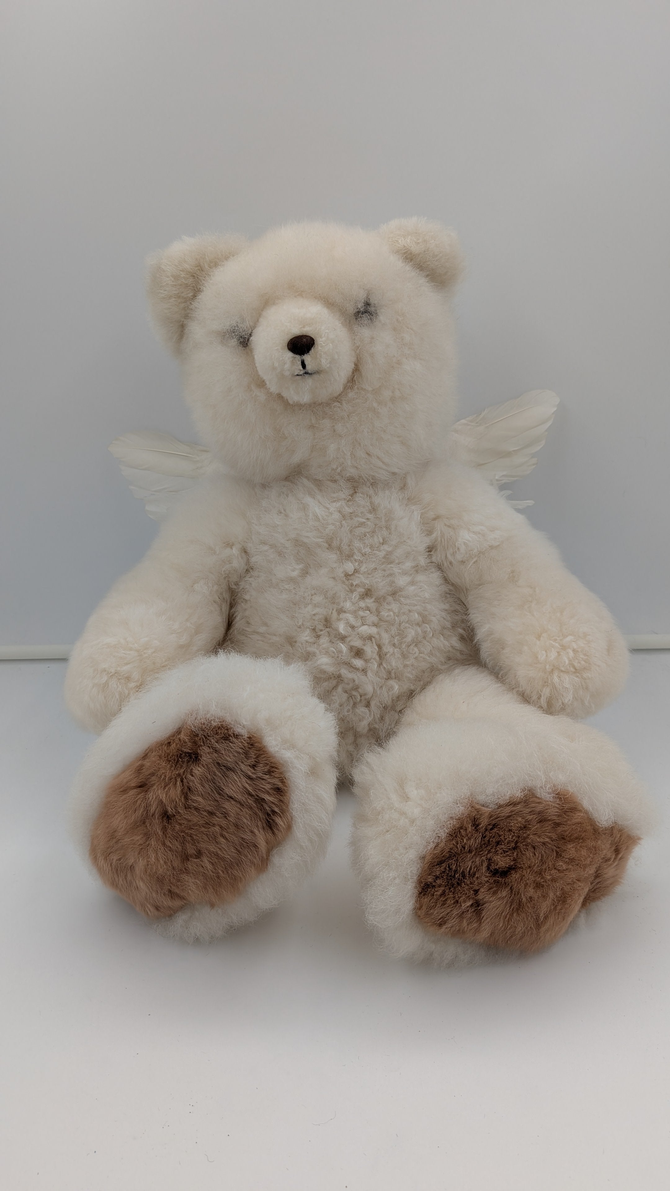 A fluffy cream-colored plush teddy bear with small angel wings and brown paw pads, sitting against a plain light gray background.