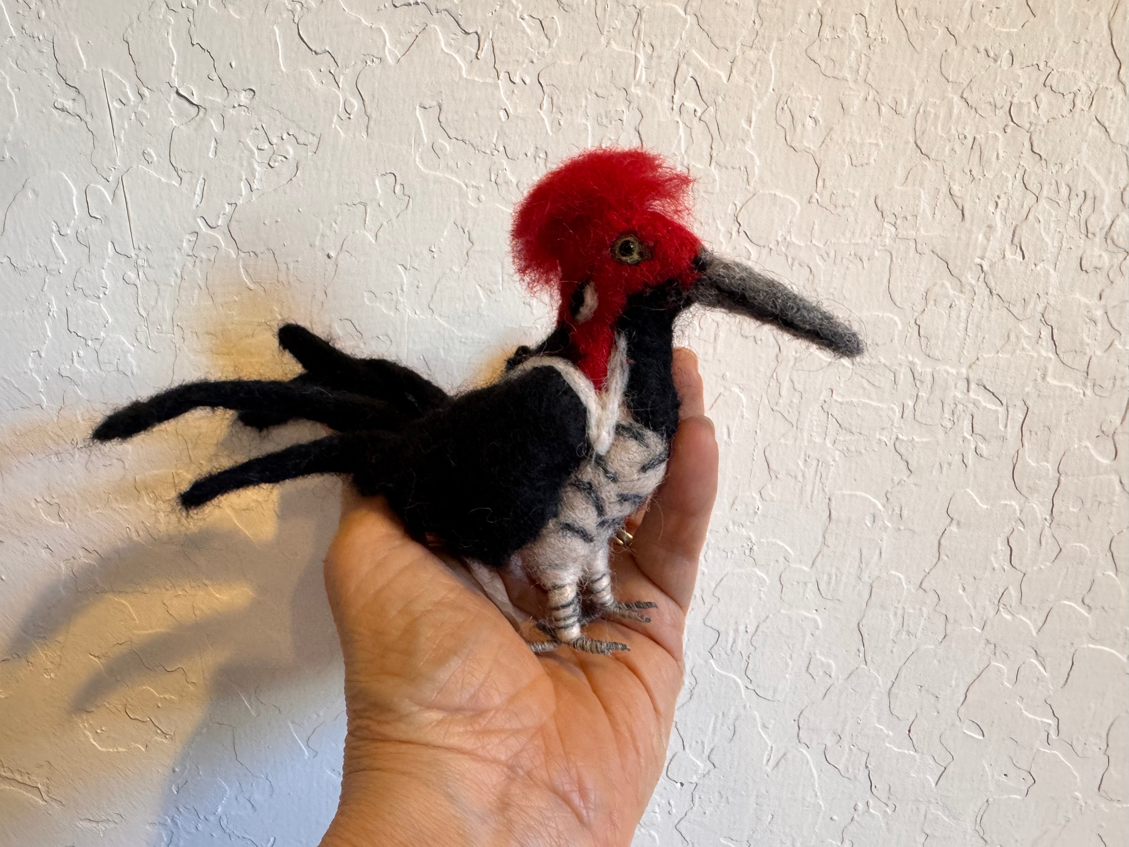 Hand holding a small woodpecker bird decor with a red head against a textured white wall.