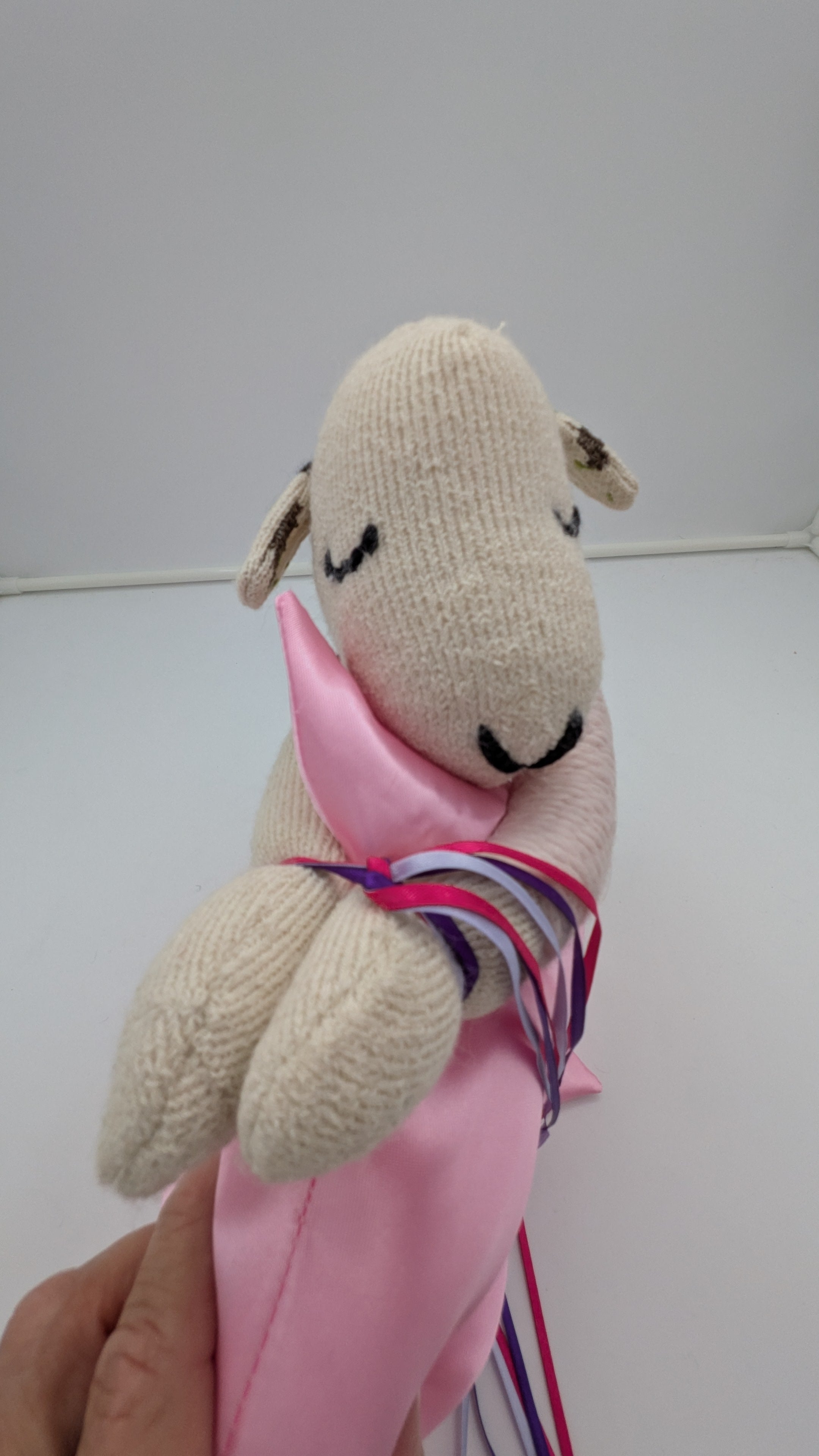 A handmade knit sheep plush toy being held by a hand. The sheep is cream-colored with black embroidered eyes and nose, wrapped in a pink ribbon with purple and blue stripes.