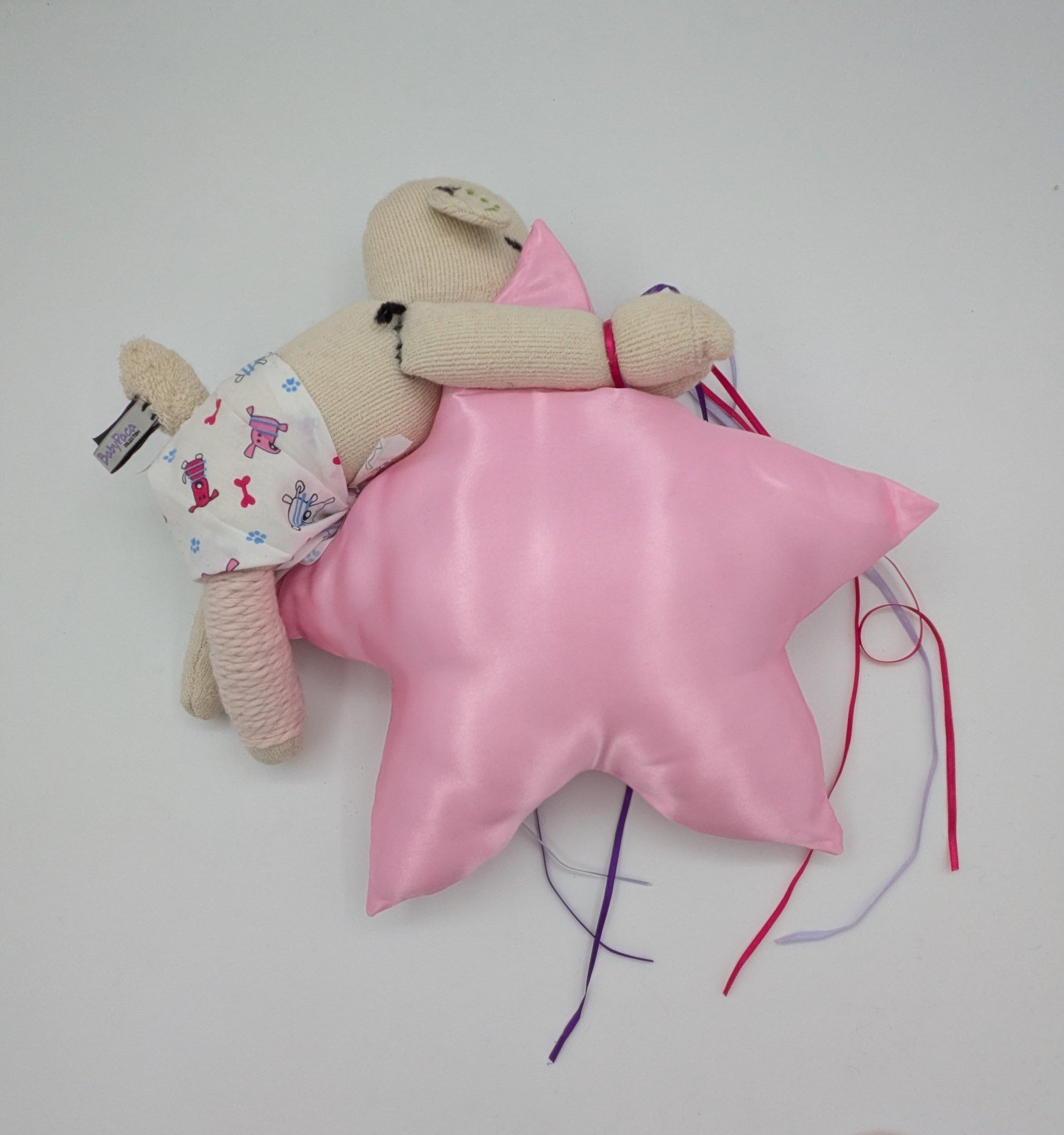 A small beige plush doll wearing a white floral outfit lies on top of a shiny pink star-shaped pillow with colorful thread tails, photographed on a plain white surface.