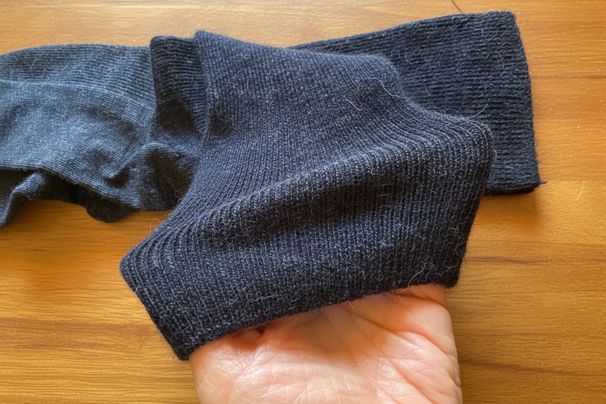 A close-up of a dark gray ribbed knit sock being held by a hand against a tan cardboard background.