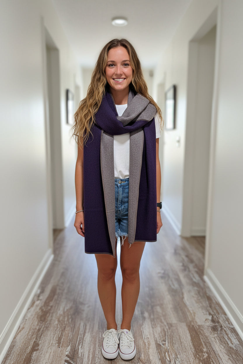 A smiling woman stands indoors wearing a folded dark purple and heather gray knit scarf or wrap around her shoulders, paired with a white t-shirt, denim shorts, and white sneakers.