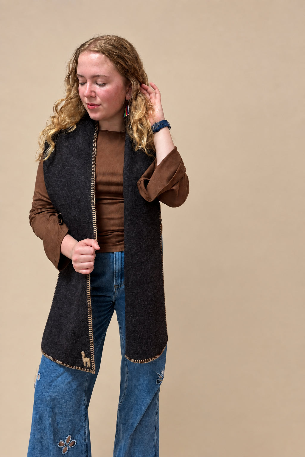 A women modeling a dark brown alpaca wool scarf with gold trim and a small alpaca patch on the hem, styled with blue jeans against a solid beige background.