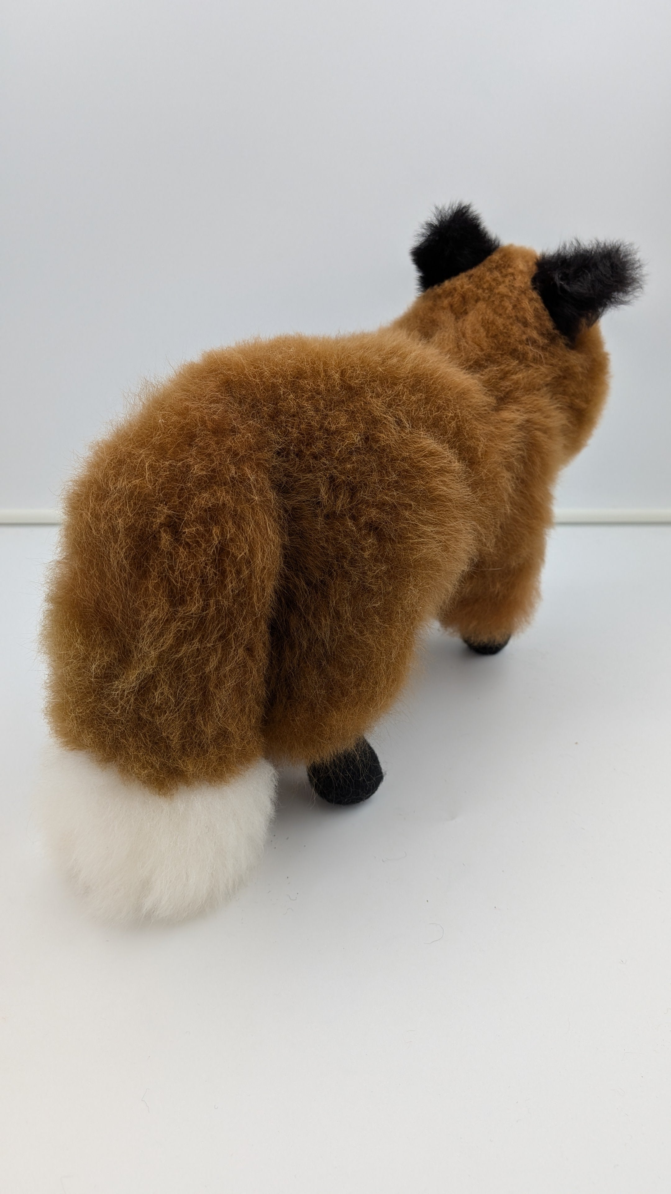 A plush toy fox shown from the back, featuring brown fur, a white-tipped tail, and black ears and paws, photographed against a plain white background.