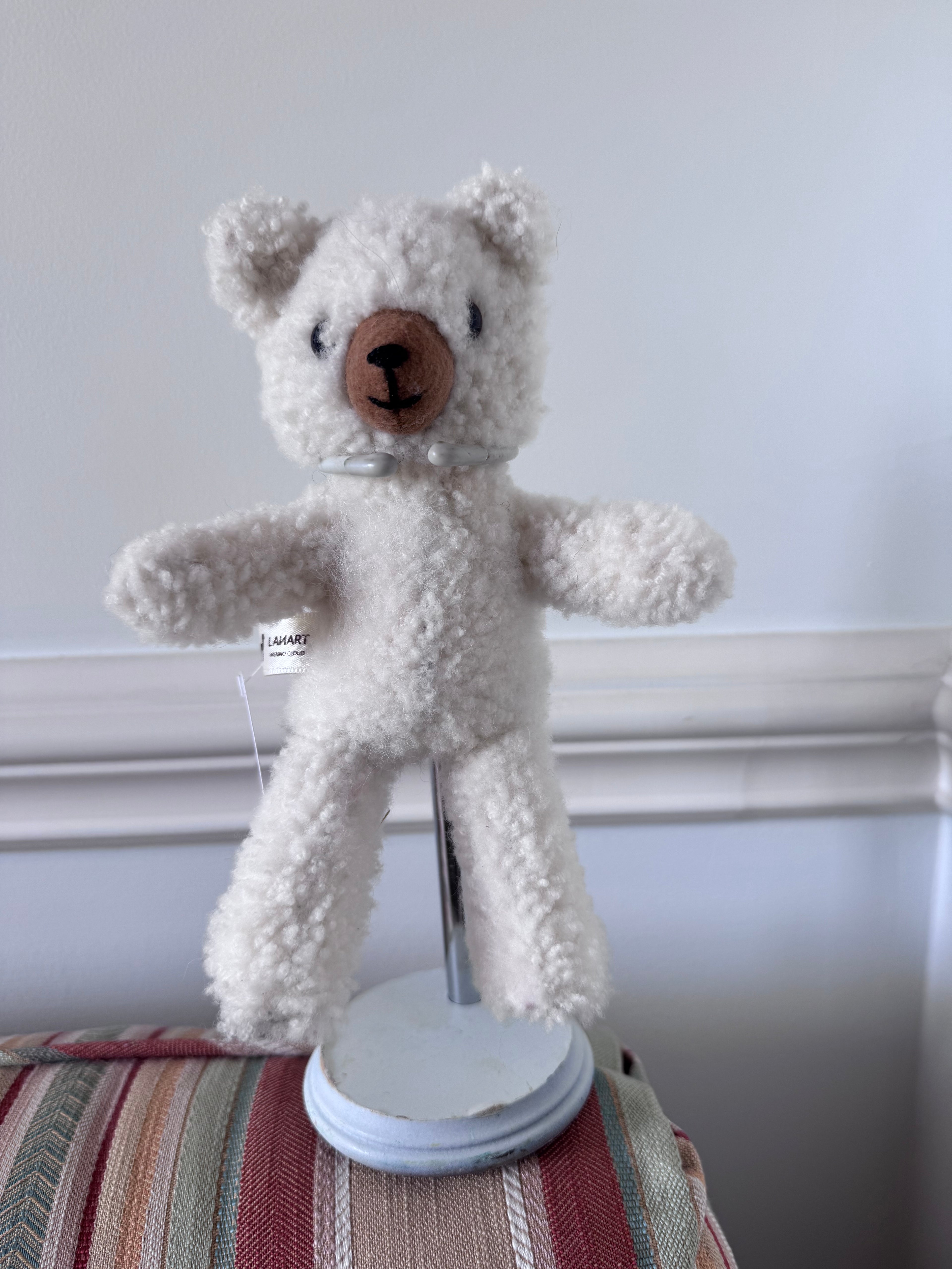 A small cream-colored teddy bear with a brown snout and black stitched eyes, displayed on a white stand on top of a striped cushion against a white wall.