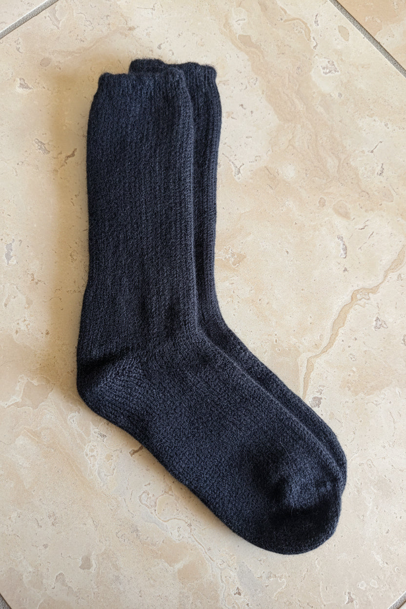 A pair of dark charcoal gray knit socks laid flat on a brown cardboard box, with a gray stone-tile floor visible in the background.