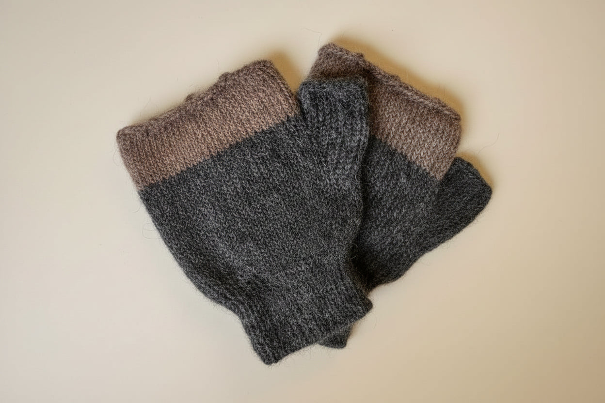 A pair of folded knit fingerless gloves in heathered brown and dark gray, with a white circular brand tag attached, laid on a light off-white textured fabric background.
