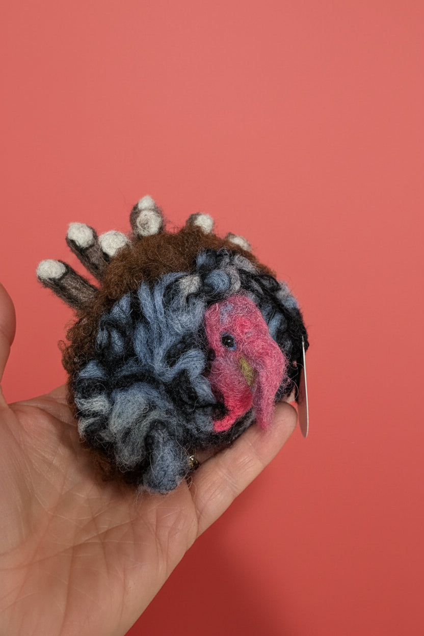 A hand holding a small felted wool ornament shaped like a turkey, with gray and black mottled feathers, a pink head, and brown body, photographed indoors against a white surface.