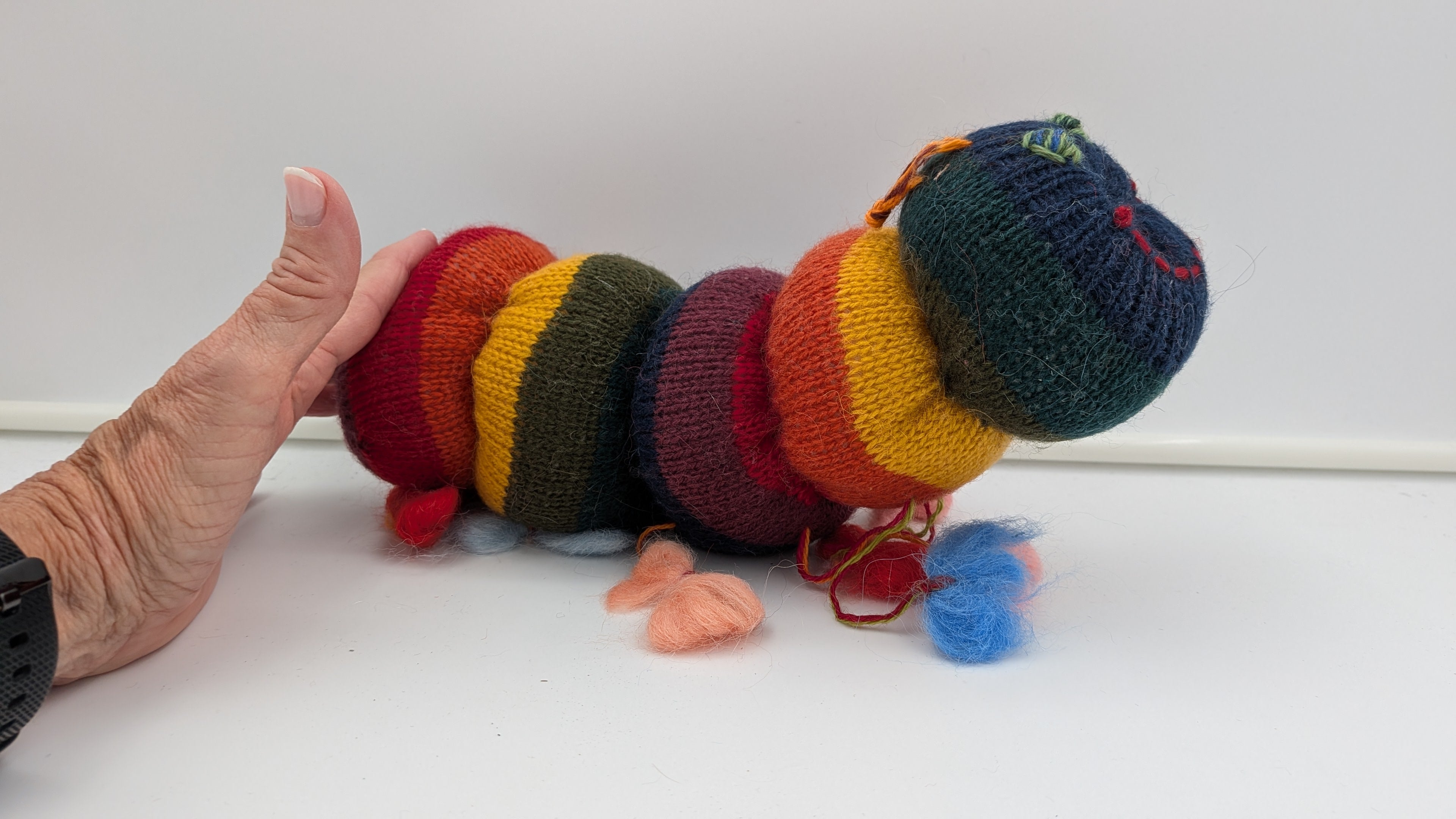 Colorful knitted caterpillar toy on a white surface with a hand pointing at it.