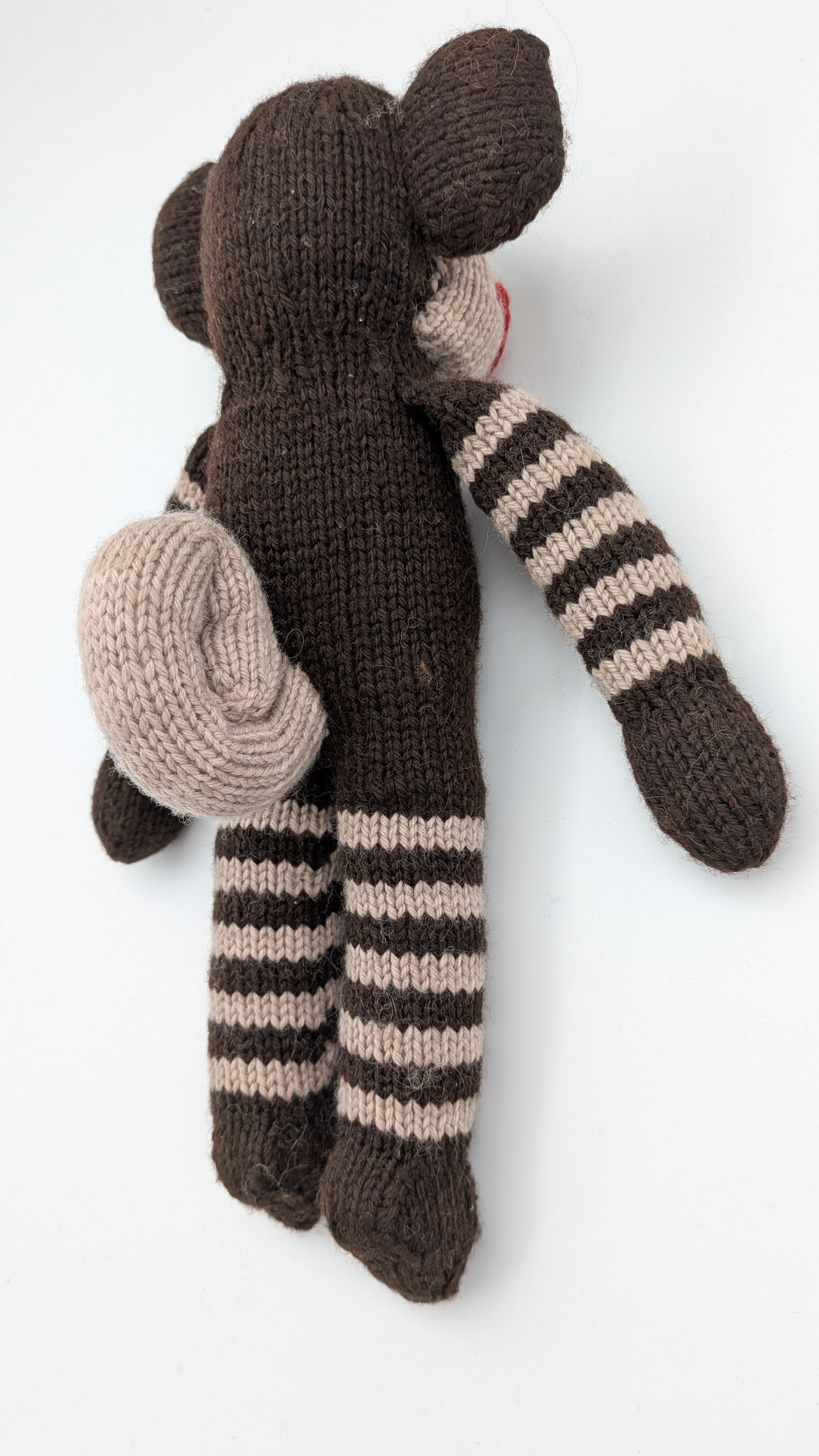 A handmade knitted monkey doll lying on its back, featuring a dark brown body, beige and brown striped arms and legs, and a beige hat, photographed on a white background.
