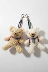 Two small teddy bear keychains made of textured plush fabric, one in beige and one in cream, each with a colorful braided strap and silver lobster clasp, displayed on a plain white surface.