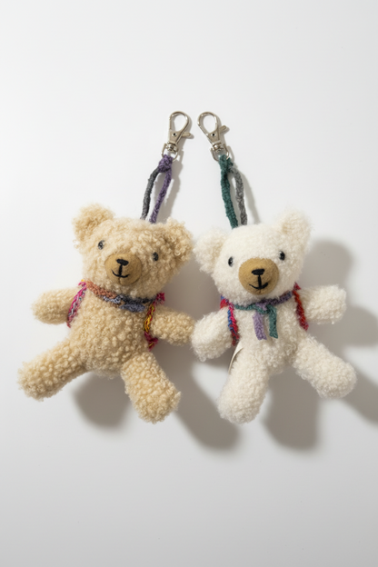 Two small teddy bear keychains made of textured plush fabric, one in beige and one in cream, each with a colorful braided strap and silver lobster clasp, displayed on a plain white surface.