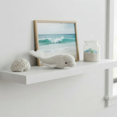 Lanart White alpaca fiber beluga whale toy on a shelf with a framed picture of waves and a small jar.