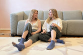 Two women sitting on a beige patterned rug in front of a textured gray sofa, wearing matching blue crew socks with multicolor accents on the toes and heels.