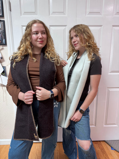 Two women modeling long knit cardigans in an indoor setting with a white paneled door behind them. The woman on the left wears a dark brown cardigan over a brown top and blue jeans, while the woman on the right wears a light sage green cardigan over a black top and ripped blue jeans.