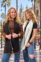 Two women standing outdoors on a palm-tree-lined street, each wearing a lanart scarves. The woman on the left has long curly blonde hair and wears a brown top with blue jeans.