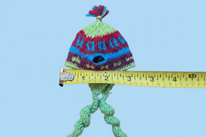 A handmade multicolor knit hat with a pom-pom next to a yellow measuring tape on a light surface.