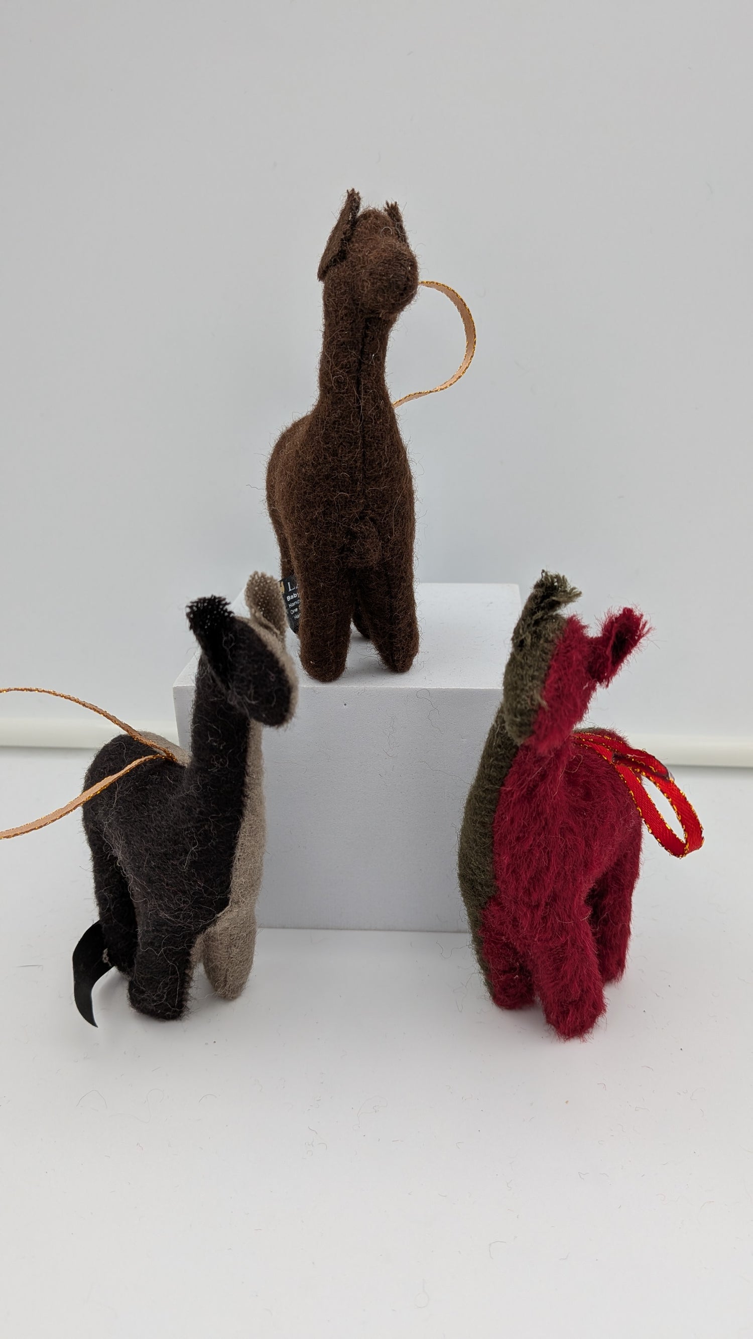 Three small felt llama or alpaca-shaped ornaments in brown, black, and red, each with a looped string and hanging loop, displayed on a white block against a light gray background.