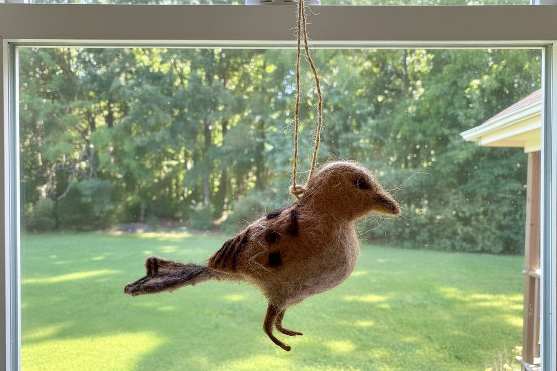 A felted wool bird ornament with brown, gray, and black coloring, suspended by a rope from a circular hanging piece, photographed outdoors in a backyard setting.