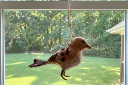 A felted wool bird ornament with brown, gray, and black coloring, suspended by a rope from a circular hanging piece, photographed outdoors in a backyard setting.