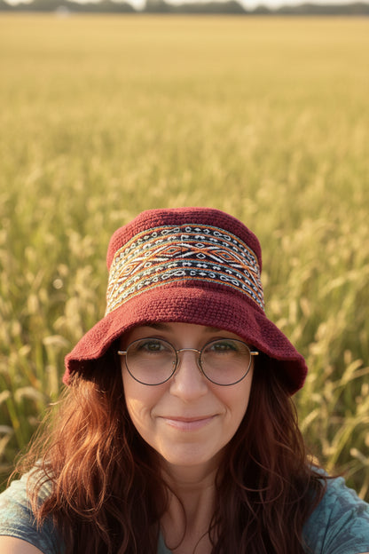 A woman wearing a burgundy knit bucket hat with a colorful geometric patterned band, photographed in an indoor setting with a painting and striped sofa in the background.