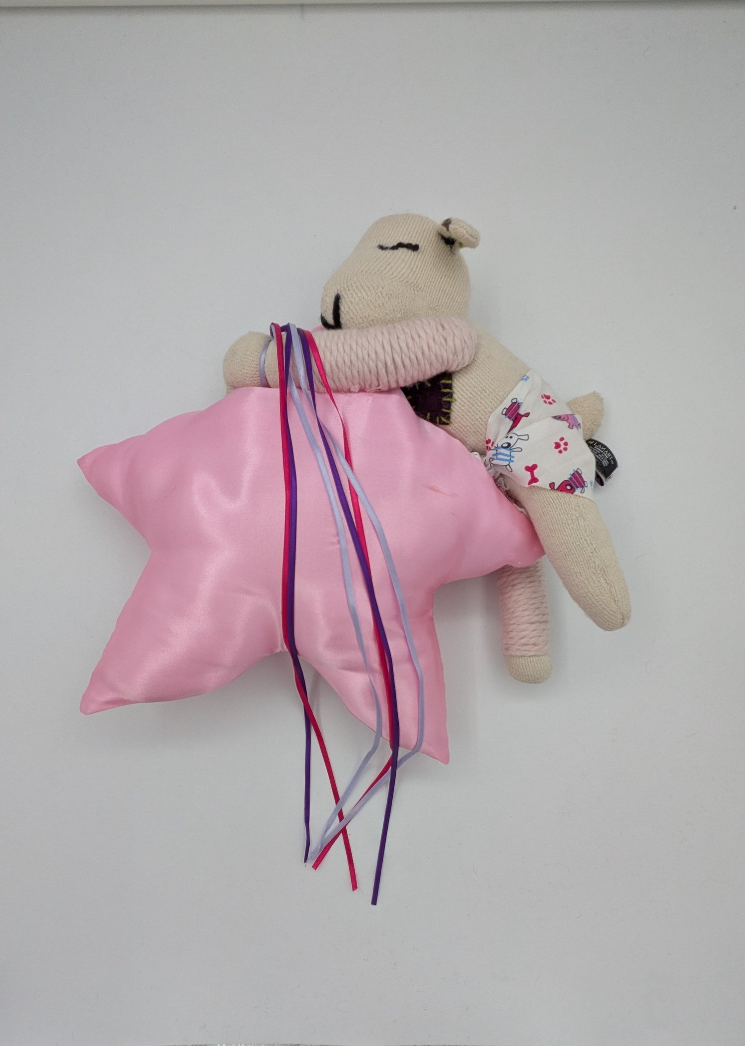 A small beige plush toy or stuffed animal lying on a white surface, wrapped in a shiny pink satin cloth with long multicolored ribbons tied around it. The toy has embroidered facial features and a tiny floral-patterned accessory.