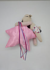 A small beige plush toy or stuffed animal lying on a white surface, wrapped in a shiny pink satin cloth with long multicolored ribbons tied around it. The toy has embroidered facial features and a tiny floral-patterned accessory.