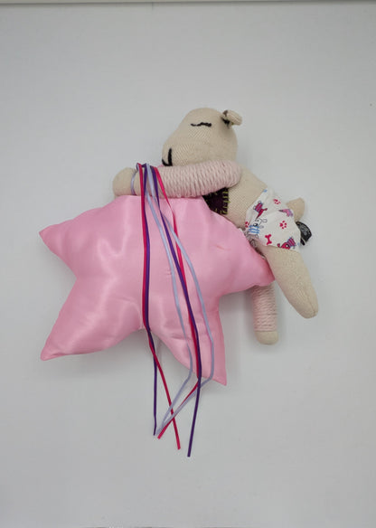 A small beige plush toy or stuffed animal lying on a white surface, wrapped in a shiny pink satin cloth with long multicolored ribbons tied around it. The toy has embroidered facial features and a tiny floral-patterned accessory.