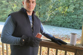 A man wearing a black fleece vest, black gloves, and sunglasses stands on a wooden deck outdoors. He is holding a small brown rectangular item, with a white insulated tumbler resting on the railing beside him.