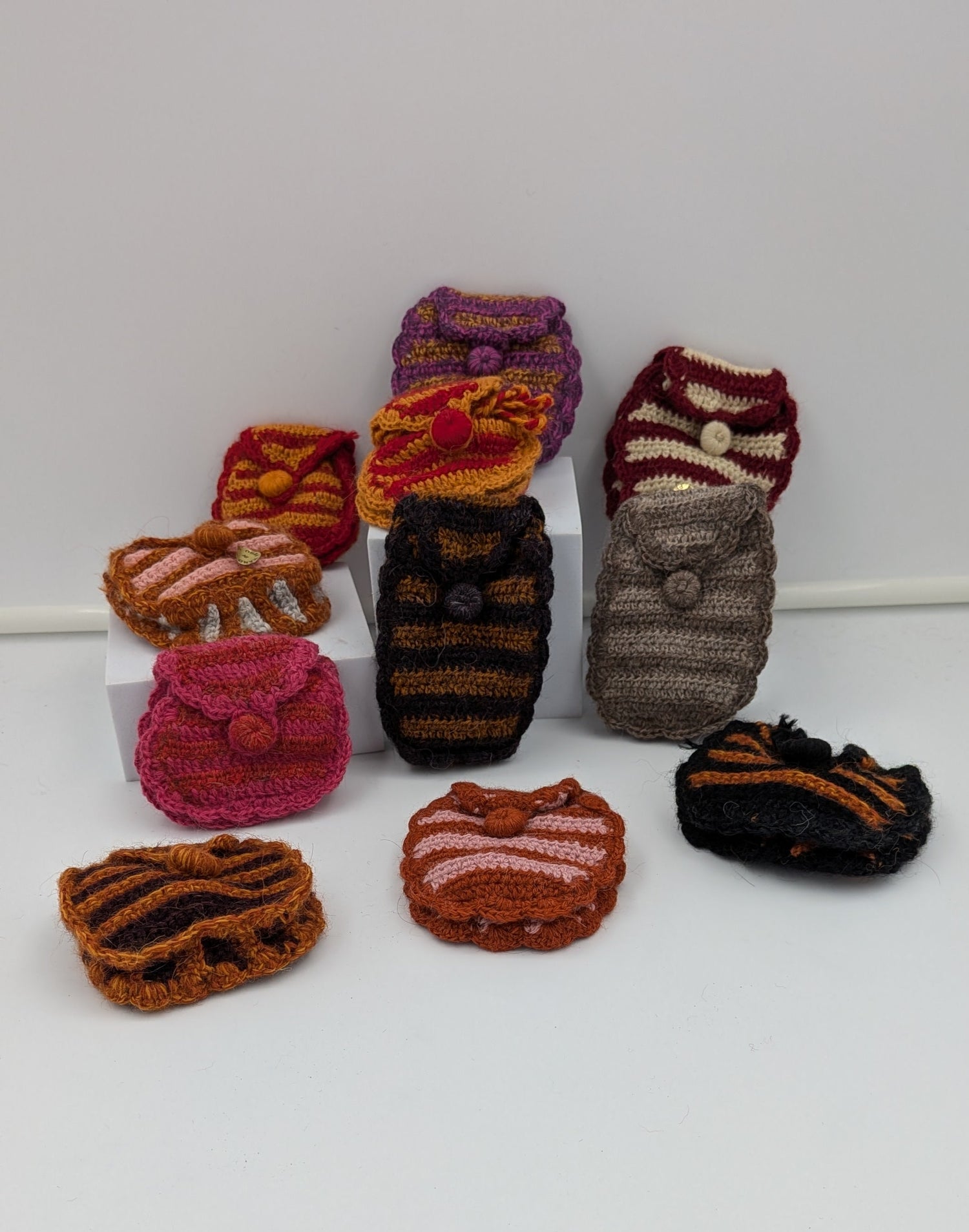 A group of small hand-knit or crocheted pouches and small accessories displayed on a white surface against a white background. The items include striped and solid knit pieces in pink, brown, orange, red, gray, and black.
