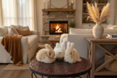 Three fluffy faux-fur bunny toys in beige, white, and cream, arranged on a round red patterned table in front of a brick wall.
