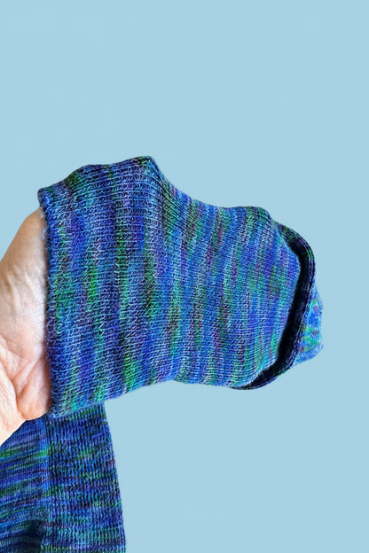 A close-up of a hand holding a folded pair of blue and green variegated knit socks or leg warmers against a cardboard box background.