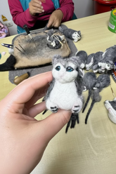 Hand holding a small felted animal toy with more toys on a table in the background.