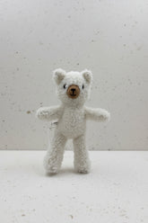 A small white teddy bear plush toy with a brown button nose and black embroidered eyes, mounted on a clear stand against a plain light gray wall.