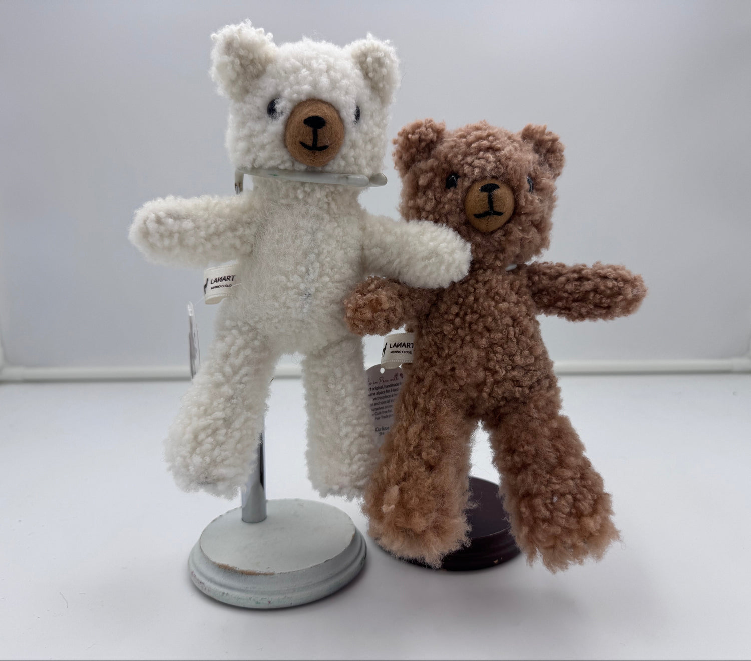 Two small teddy bears made of curly plush fabric, one cream/off-white and one medium brown, standing on display stands against a light gray studio backdrop.