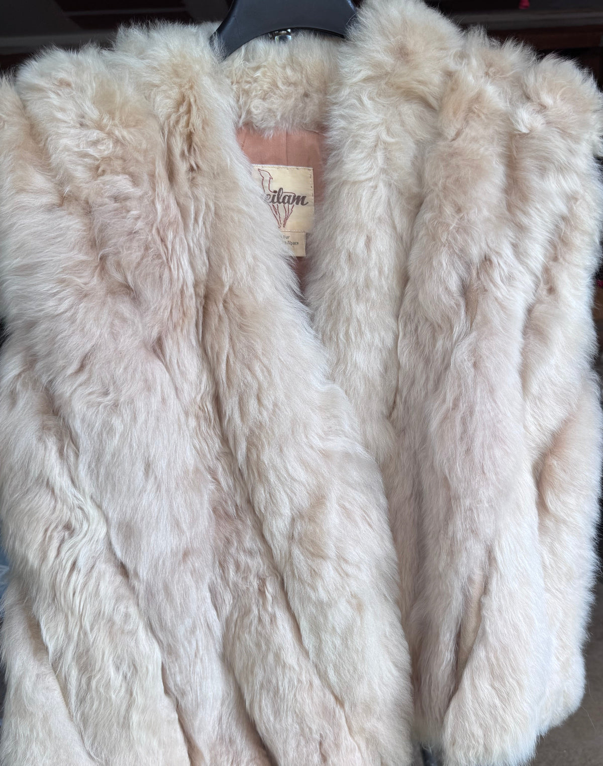 A large, fluffy fur vest in  beige color, displayed on a black hanger.
