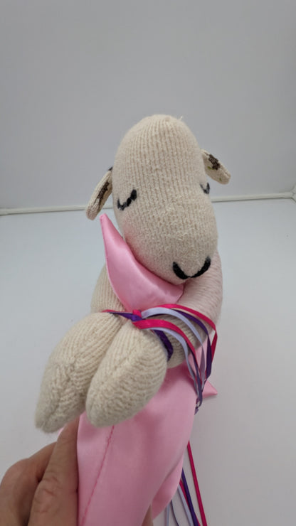 A handmade knit sheep plush toy being held by a hand. The sheep is cream-colored with black embroidered eyes and nose, wrapped in a pink ribbon with purple and blue stripes.