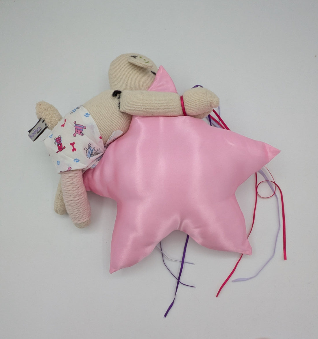 A small beige plush doll wearing a white floral outfit lies on top of a shiny pink star-shaped pillow with colorful thread tails, photographed on a plain white surface.