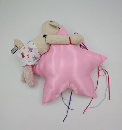 A small beige plush doll wearing a white floral outfit lies on top of a shiny pink star-shaped pillow with colorful thread tails, photographed on a plain white surface.