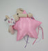 A small beige plush doll wearing a white floral outfit lies on top of a shiny pink star-shaped pillow with colorful thread tails, photographed on a plain white surface.