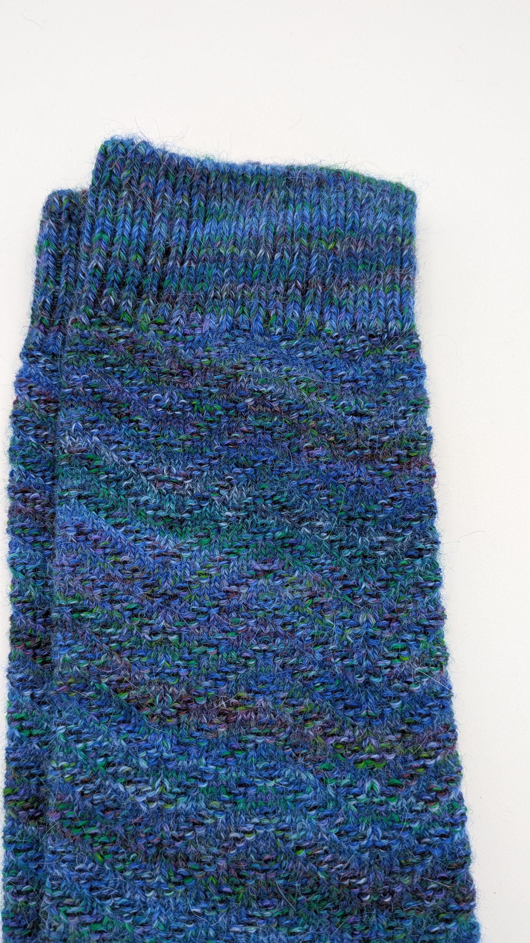 A folded pair of hand-knit socks in a variegated blue and green yarn, showing a ribbed cuff and textured knit body on a plain white background.