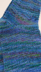 Close-up of a hand-knit sock in variegated blue, green, and purple yarn, showing the detailed knit texture.