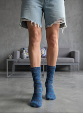 A person wearing blue and navy marled knit crew socks, a gray skirt, and a gray blazer, standing in a modern gray living room.