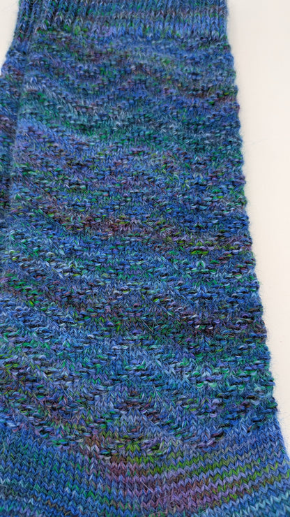 A close-up of a hand-knit sock in a variegated blue, teal, and purple yarn, laid flat on a white background.