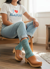 A person sitting on a wooden chair in a bright room, wearing light blue jeans, a white graphic T-shirt, and teal knit clogs with tan wood heels. The person is cropped from the shoulders down, and the background shows a window, floor, and rug.