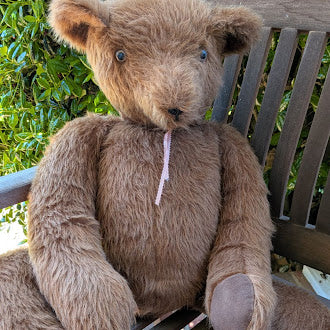 Brown teddy bear sitting on a wooden bench with greenery in the background