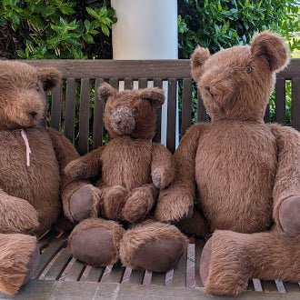 Three brown teddy bears sitting on a wooden bench outdoors.