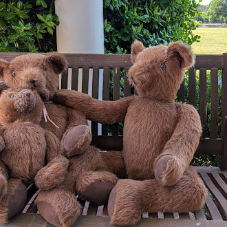 Three brown teddy bears sitting on a wooden bench outdoors.
